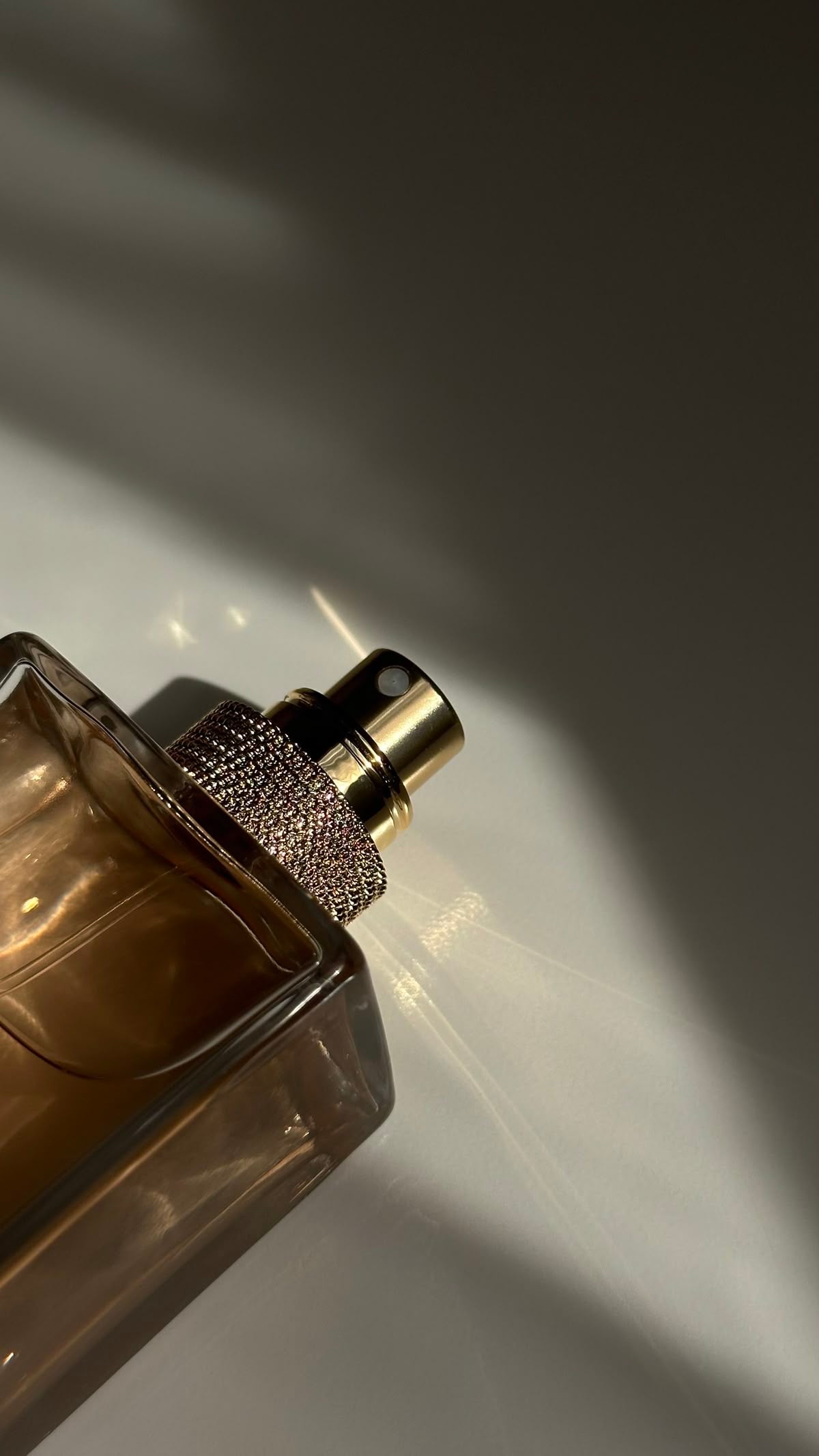 Frasco de perfume con vaporizador dorado y textura de cristales, iluminado por la luz natural sobre fondo neutro, evocando elegancia y sutileza para la estética ROUCLÊ