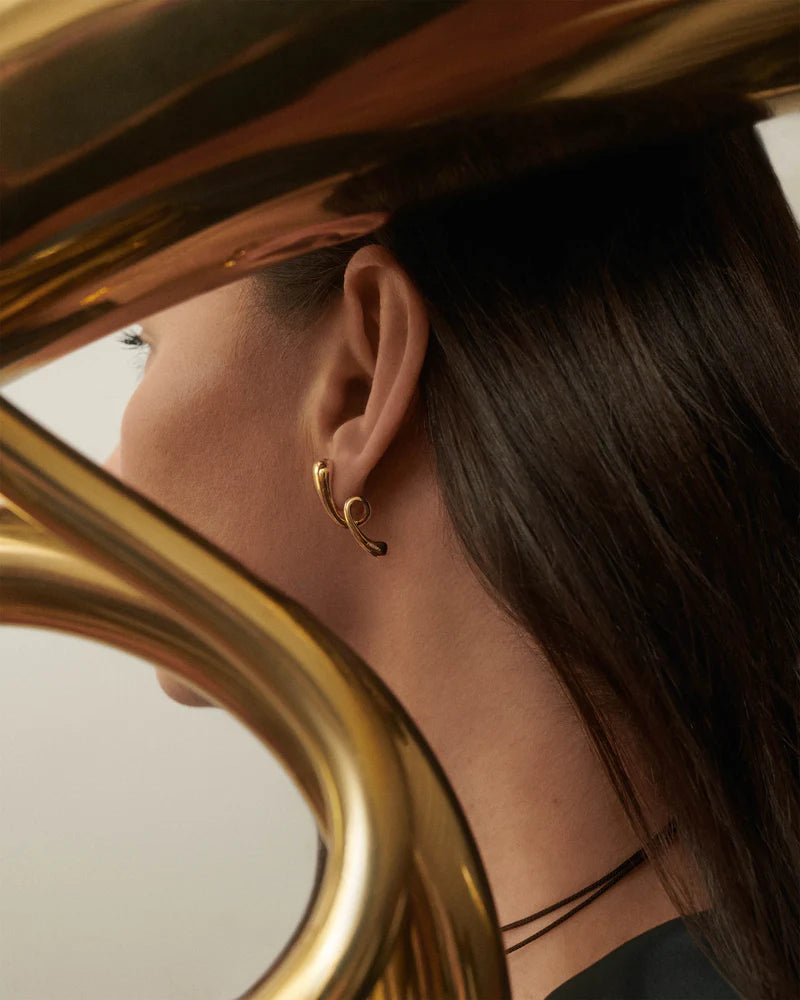 Pendientes Orbit de PDPAOLA en plata bañada en oro, fotografiados en la oreja de una mujer, con fondo de un instrumento musical dorado, resaltando el diseño escultórico de gota