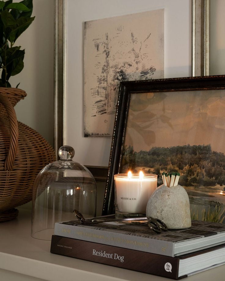 Vela perfumada encendida junto a objetos decorativos, libros de diseño y obras de arte enmarcadas, creando un ambiente elegante y acogedor.