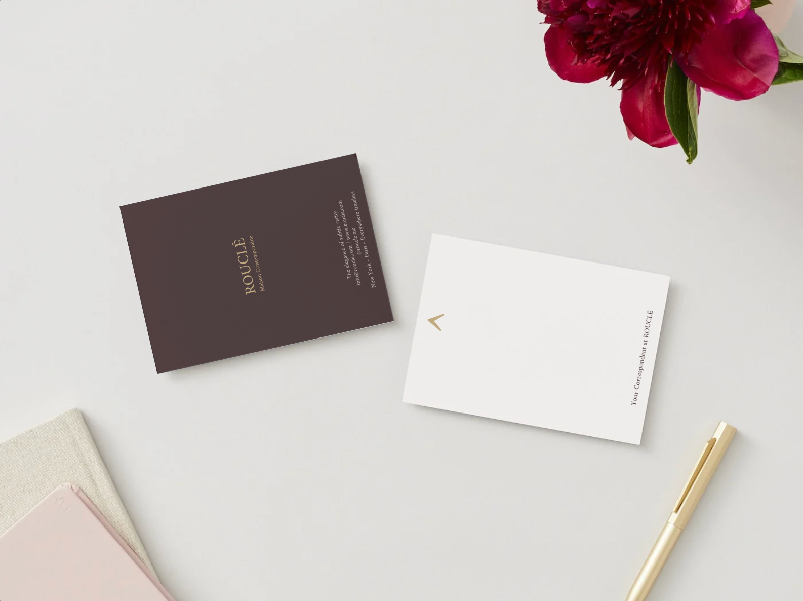 Tarjetas editoriales de ROUCLÊ sobre mesa clara, con flor roja y bolígrafo dorado. Una tarjeta marrón con logo y lema, y otra blanca con símbolo ^ y la firma Your Correspondent at ROUCLÊ.