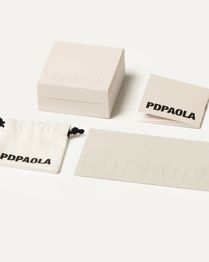 Estuche y funda de tela de la marca PDPAOLA con logotipo visible, en presentación de joyería elegante.