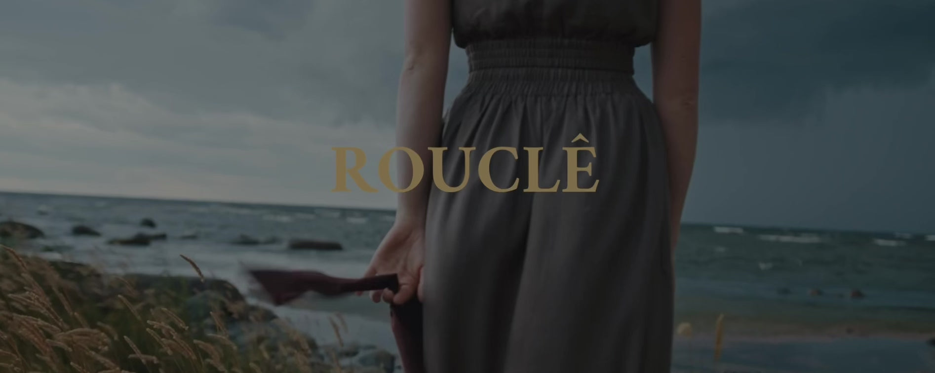 Video editorial de ROUCLÊ: mujer en la orilla con vestido vaporoso, sosteniendo un sombrero, mientras el mar se funde con el logo dorado.